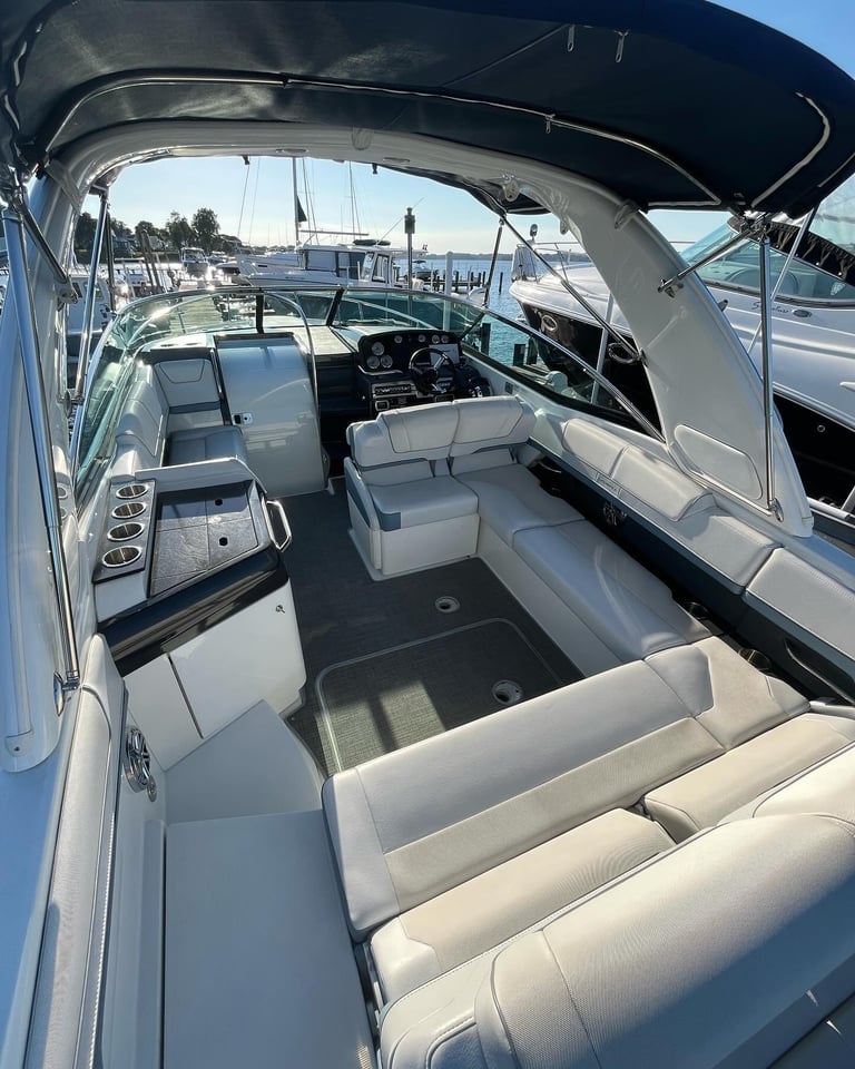 Interior view of a white speedboat with cream seating, steering wheel, and control dashboard, docked at a marina with sailboats visible in the background