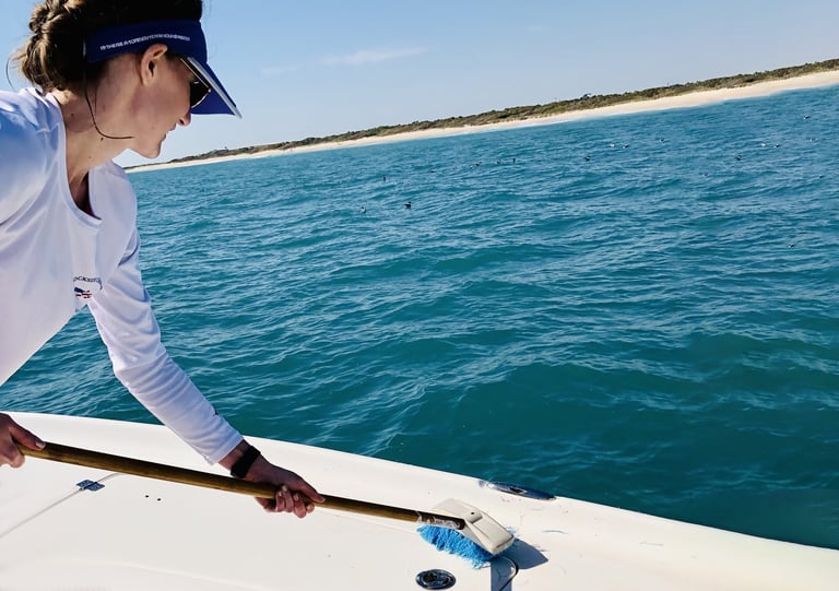 Person in white long-sleeve shirt and blue visor leaning over boat railing, looking at turquoise ocean water near sandy coastline