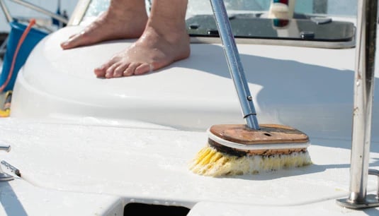 Person cleaning boat deck with long-handled brush, barefoot on white surface near metal rail