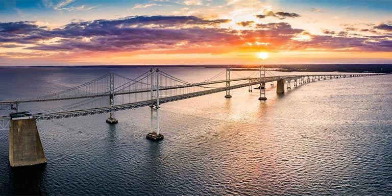 Aerial view of a long bridge spanning across blue water at sunset with vibrant orange and purple sky